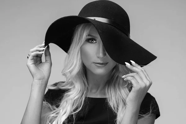 Young blond woman in big black stylish hat and black dress is looking into the frame. She has a clean and healthy skin, a serious look