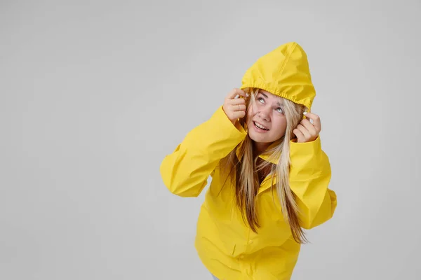 Ung kvinna blondin i en gul regnrock, med en luva på huvudet bakgrund hudar från ett regn på en grå bakgrund — Stockfoto