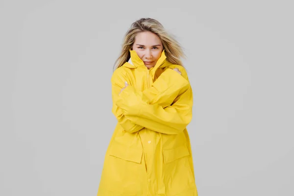 灰色の背景に黄色のレインコートで若い金髪女性が保温しようと雨の中から冷凍 — ストック写真