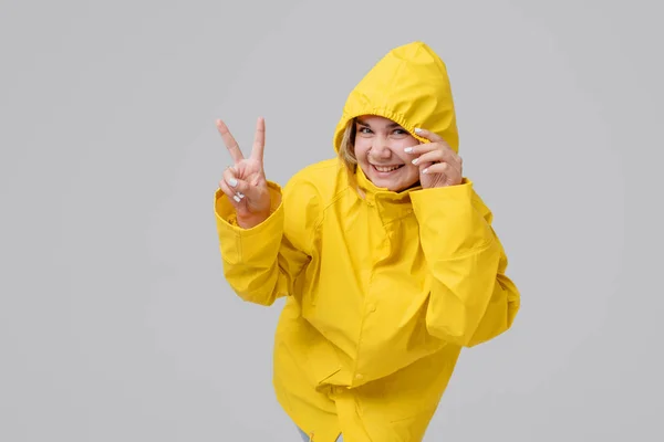 Giovane donna bionda in un impermeabile giallo, con un cappuccio su uno sfondo testa si nasconde da una pioggia su uno sfondo grigio — Foto Stock