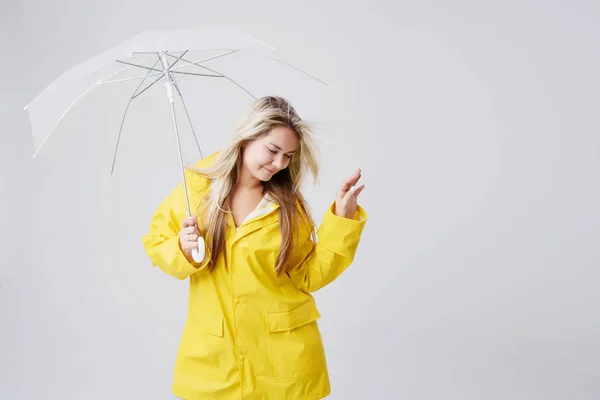 Blonde woman wearing yellow raincoat holding transparent umbrella checking weather if it is raining. Protected against rain and hurricane