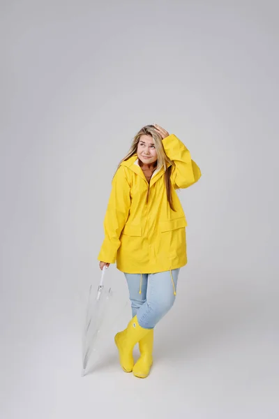 Mujer rubia vistiendo impermeable amarillo sosteniendo paraguas transparente comprobando el clima si está lloviendo. Protegido contra la lluvia y el huracán —  Fotos de Stock