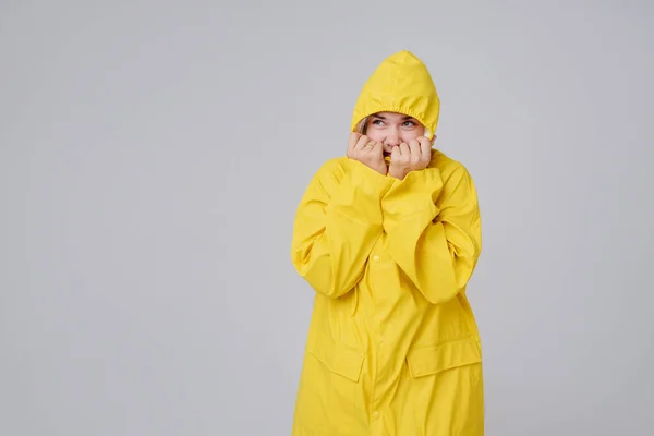 Mulher jovem loira em uma capa de chuva amarela, com um capuz no fundo da cabeça se esconde de uma chuva em um fundo cinza — Fotografia de Stock