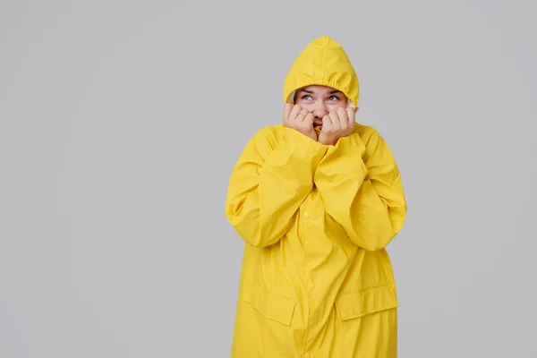 Ξανθιά νεαρή γυναίκα σε ένα κίτρινο αδιάβροχο, με μια κουκούλα στο κεφάλι φόντο κρύβει από μια βροχή σε γκρίζο φόντο — Φωτογραφία Αρχείου