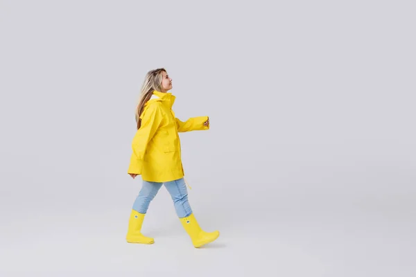 黄色いレインコートを着て、白い背景の上の黄色のゴム製ロングブーツで金髪の若い女性。雨の後のステップは喜んで、水たまりを飛び越えて — ストック写真