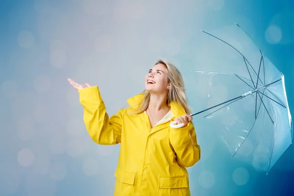 Blond kvinna klädd i gul regnrock innehar transparent paraply kontrollera vädret om det regnar. Skyddad mot regn- och orkansäsongen — Stockfoto