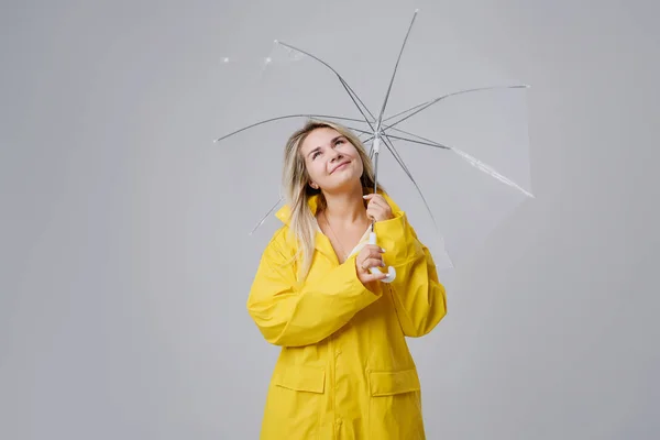 Szőke nő visel a sárga esőkabátot gazdaság átlátszó esernyő, időjárás ellenőrzése, ha esik az eső. Eső és hurrikán ellen védett — Stock Fotó