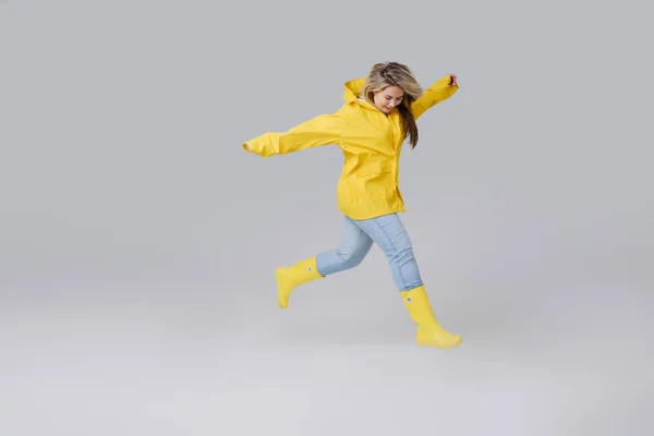 Giovane donna bionda in un impermeabile giallo e stivali al ginocchio in gomma gialla su uno sfondo bianco. Volentieri passi dopo una pioggia e salti sopra attraverso pozzanghere — Foto Stock
