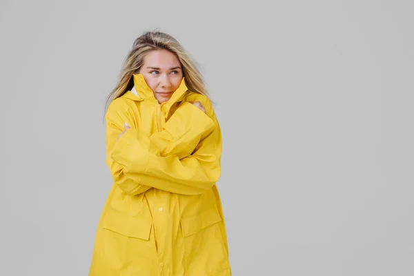 Young blonde woman in a yellow raincoat on a gray background is frozen from the rain and trying to keep warm