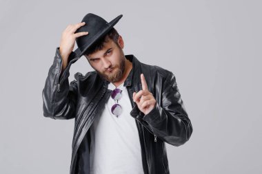 Bir şık siyah deri ceket ve gri bir arka plan üzerine şapka sakallı genç bir neşeli adam. İyi haber gülümsüyor. Bir başparmak havaya