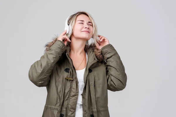 一个穿着秋季保暖夹克的年轻女孩在灰色背景上收听无线耳机上的音乐 — 图库照片
