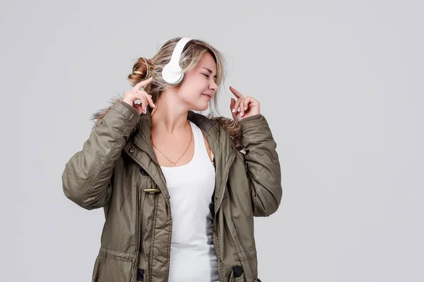 一个穿着秋季保暖夹克的年轻女孩在灰色背景上收听无线耳机上的音乐 — 图库照片
