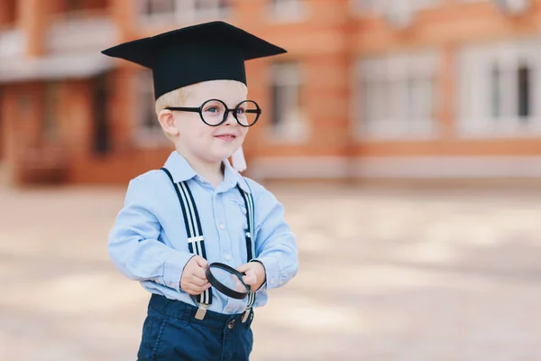 スマートの小さな男の子に眼鏡 学術の帽子と眼鏡学校の裏庭に立っています 彼の手で虫眼鏡を保持します — ストック写真