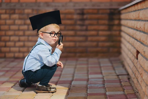 眼鏡や学校のレンガの壁に対して立っている学術の帽子の少年 虫眼鏡を保持し 環境を研究 — ストック写真