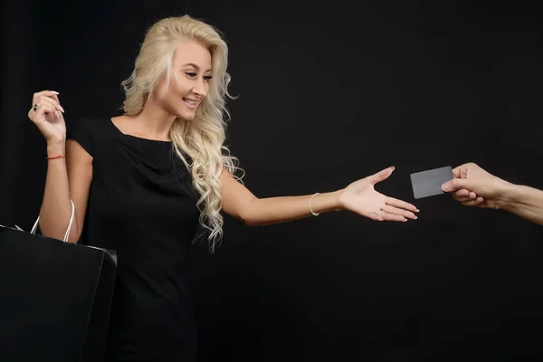 Young blond woman with long hair in a stylish black dress and black package pulls a hand to a credit card on a black background