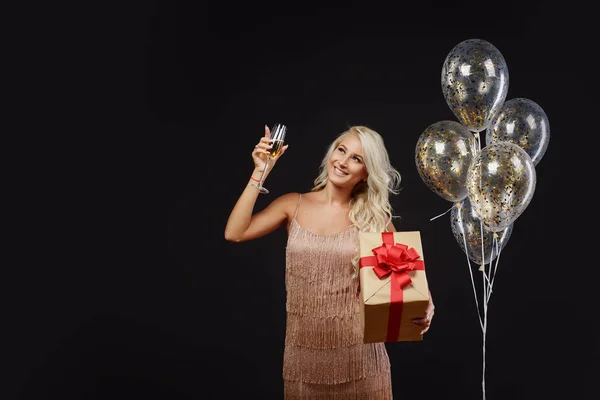 Joyful young woman in luxury dresses celebrating birthday or Christmas party with confetti on black background.Having fun and smiling. Holding present, gift, golden balloons and glass of champagne
