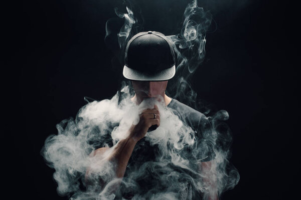 Vape man. Portrait of a handsome young white guy in a modern black cap vaping and letting off puffs of steam from an electronic cigarette