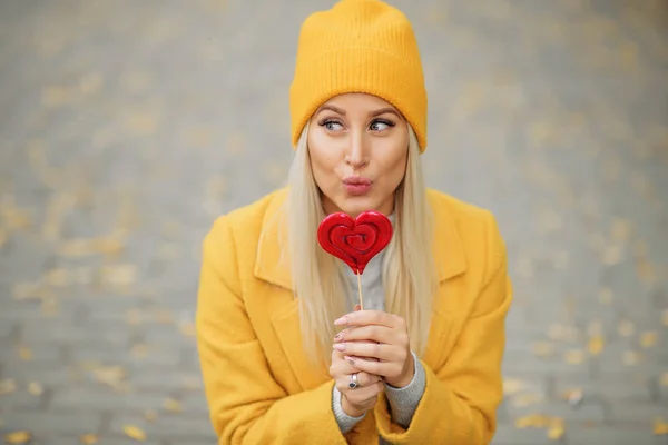 Fashion portrait pretty sweet young woman in yellow coat having fun with red lollipop heart over autumn street background