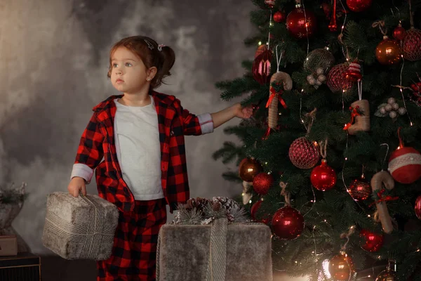 愉快的小微笑的女孩在红色睡衣与圣诞礼物箱子在客厅在家居装饰 — 图库照片