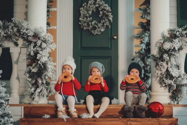 A portrait of children sitting on the porch of their house together, Eat bagels. Merry Christmas, happy New Year. Yard with a christmas tree, lights and decorations. Miracle time.