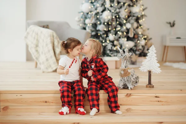 Two little sisters in red pajamas sit in the living room on Christmas morning and eat sweets. Family party, hugs and kisses