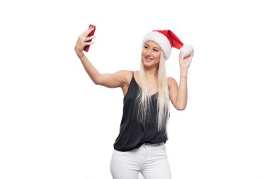 Gülümseyen mutlu sarışın kadın siyah bluz ve Noel şapka selfie kırmızı smartphone kamera üzerinden yapmak Noel Baba beyaz arka plan izole. Satış konsepti