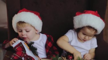 Genç çocuk, kardeşi ve kız kardeşi, Noel Baba şapkaları Noel ağacının altına oturma odasında oturmak ve hediyeleri açalım. Oda ışık ampuller ile geleneksel dekorasyon. Aile kavramı