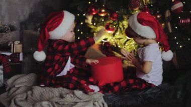 Genç çocuk, kardeşi ve kız kardeşi, Noel Baba şapkaları Noel ağacının altına oturma odasında oturmak ve hediyeleri açalım. Oda ışık ampuller ile geleneksel dekorasyon. Aile kavramı