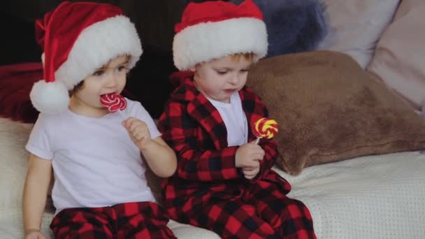 サンタ クロースの帽子にクリスマス ツリーの下のリビング ルームで座っている若い子供 兄と妹とオープン プレゼントしお菓子を食べる 家族の概念 — ストック動画