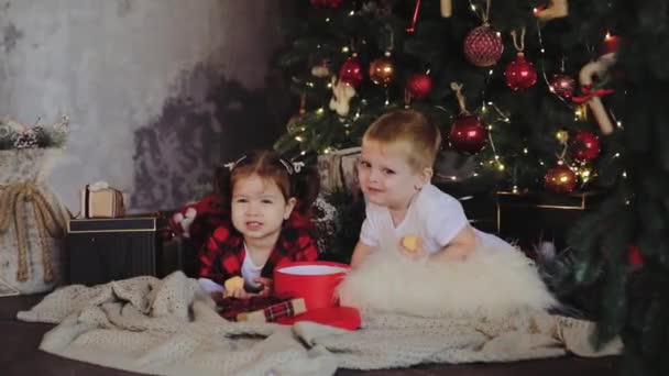 若い子供 兄と妹 サンタ クロースの帽子にクリスマス ツリーの下のリビング ルームに座っているし プレゼントを開きます 電球が付いている部屋の伝統的な装飾 家族の概念 — ストック動画
