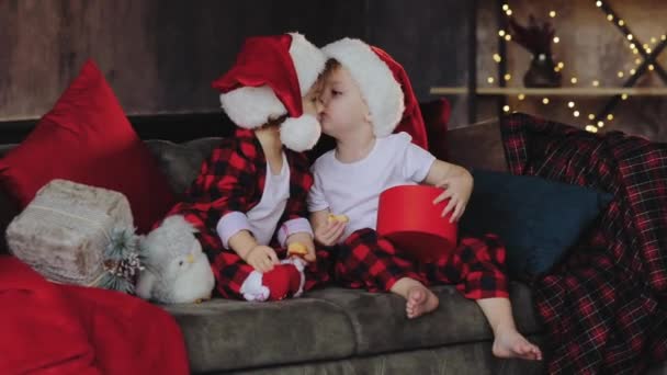 孩子们 戴着圣诞老人的帽子坐在圣诞树下的客厅里 打开礼物 房间的传统装饰 有灯泡 家庭理念 — 图库视频影像