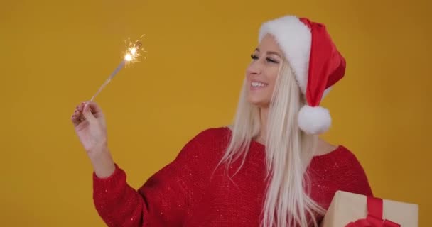 サンタ クロースの帽子の若いブロンドの女性は 花火と驚きのギフト ボックスに保持しています クリスマス パーティー 黄色の背景で楽しくダンス — ストック動画
