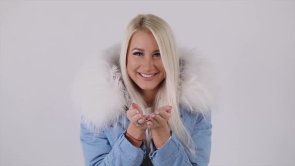 毛皮フード付きコートで若い金髪女性は願う 白い背景の上の彼女の手のひらから雪を吹く — ストック動画