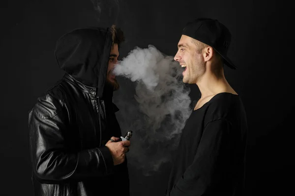 Bad habit, smoking in a public place. Two Young man blowing smoke to join it in one cloud at black studio background. Friends and vape addiction concept.