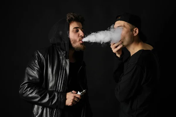 Bad habit, smoking in a public place. Two Young man blowing smoke to join it in one cloud at black studio background. Friends and vape addiction concept.
