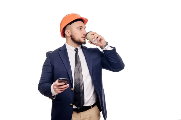 Business portrait of surprised contractor and developer, talking about phone. Businessman in hard hat with cup of coffee standing over white background. News and coffee brake concept
