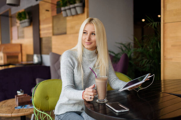 Young blond Woman using Tablet and Drinking Coffee. Relaxing in Cozy cafe, Sit on the Chair and listening with pleasure in headphones an audio book or music