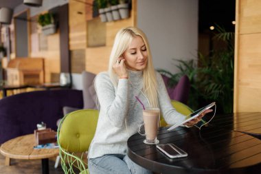 Tablet ve içme kahve kullanan genç sarışın kadın. Rahat kafede dinlendirici, sandalye ve kulaklık zevki bir sesli kitap veya müzik dinlerken oturup