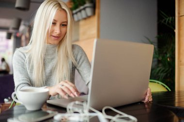 Kendine güvenen genç kadın rahat kıyafet yaratıcı ofis veya kafe ve woking otururken dizüstü bilgisayarda çalışma içinde. Bir fincan kahve tutarak ve gülümseyen 