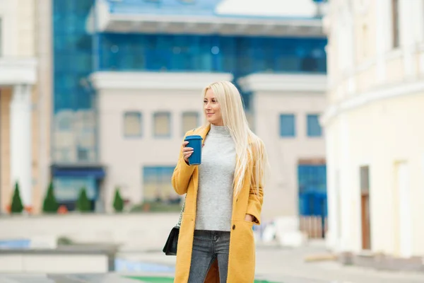 Coffee on the go. Beautiful young blond woman in bright yellow coat holding coffee cup and smiling while walking along the street