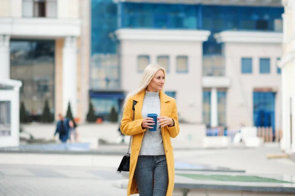 Coffee on the go. Beautiful young blond woman in bright yellow coat holding coffee cup and smiling while walking along the street