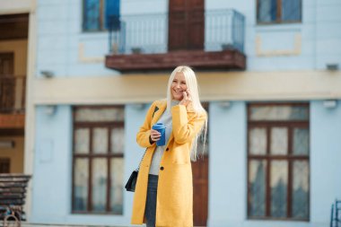 Genç sarışın bir kadın sarı parlak paltolu sokağın ortasında duran ve telefonda konuşurken. Arkadaşlar bir toplantı için bekliyor. Bir fincan kahve tutar.