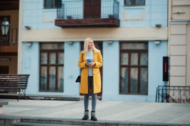 Genç sarışın bir kadın sarı parlak paltolu sokağın ortasında duran ve telefonda konuşurken. Arkadaşlar bir toplantı için bekliyor. Bir fincan kahve tutar.