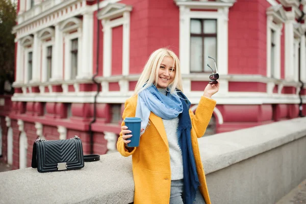 Stylish happy young blond woman in bright yellow coat holds coffee to go. Walking on the street. Portrait of a young cheerful woman at work break. Lifestyle concept