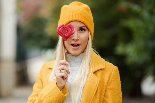 Saint Valentine\'s day concept. Fashion portrait blond young woman in yellow coat having fun with red lollipop heart over street background.