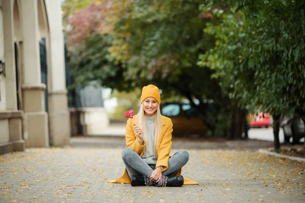 Saint Valentine\'s day concept. Fashion portrait blond young woman in yellow coat having fun with red lollipop heart over street background.