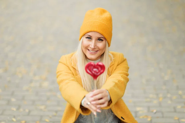 Saint Valentine\'s day concept. Fashion portrait blond young woman in yellow coat having fun with red lollipop heart over street background.