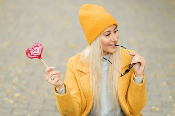 Saint Valentine\'s day concept. Fashion portrait blond young woman in yellow coat having fun with red lollipop heart over street background.