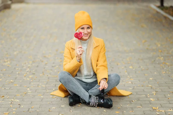 Saint Valentine\'s day concept. Fashion portrait blond young woman in yellow coat having fun with red lollipop heart over street background.