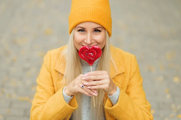 Saint Valentine\'s day concept. Fashion portrait blond young woman in yellow coat having fun with red lollipop heart over street background.